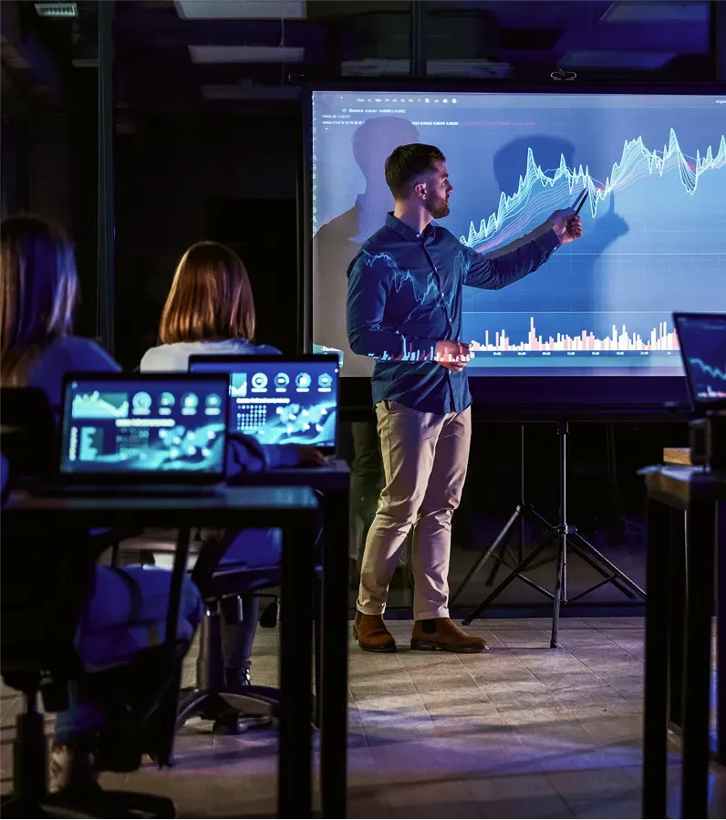 Male business trainer in a dark room presenting data on a projector, representing Top One Futures education.